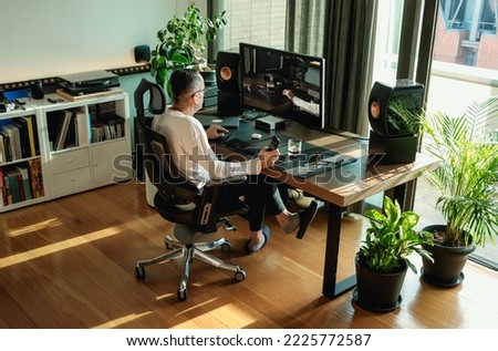 Back view of middle aged man working on his computer at home by a window at modern workplace in room decorated with green potted plants. Graphic designer working from home Royalty-Free Stock Photo #2225772587
