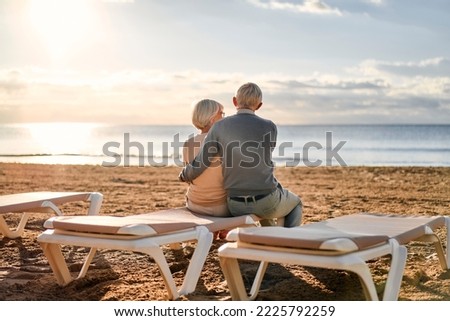 An elderly intelligent gray-haired couple in love are sitting on a sun lounger on the seashore, having a romantic time admiring the seascape.
