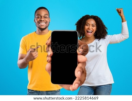 Glad emotional millennial african american couple celebrate victory with hands gesture, show big phone with empty screen, isolated on blue background, studio. Online win, new app, great ad and offer Royalty-Free Stock Photo #2225814669