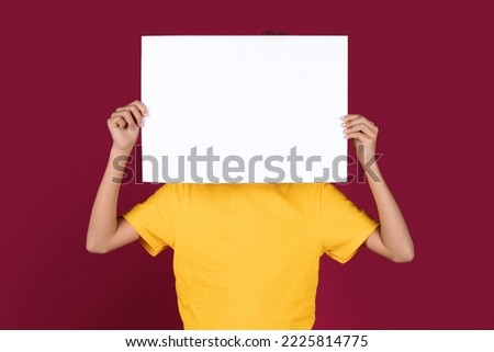 Unrecognizable african american child in yellow t-shirt hiding behind empty placard, black kid covering face with white blank advertising board, isolated on burgundy studio background, mockup Royalty-Free Stock Photo #2225814775