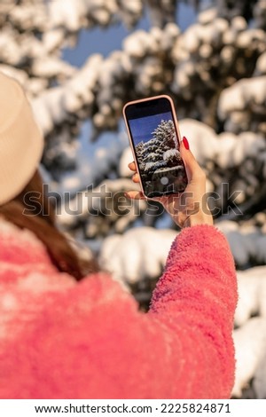 Young woman in pink coat takes pictures of snow-covered fir trees on her smartphone.Winter,technology,social media concept.Selective focus,view behind,close up.
