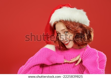 A cute girl with red hair in Santa's hat and bright pink sweater smiles happily. Red background. Christmas beauty.