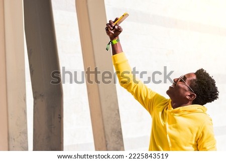 Close-up of a smiling young black man wearing a yellow sweatshirt and sunglasses taking a photo of himself in front of a white wall next to some metal columns.
