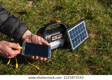 The man holds the phone in his left hand and puts it on the charger from a portable power station powered by a solar panel. A power station with a flashlight and a board powered by a solar panel. Royalty-Free Stock Photo #2225873543