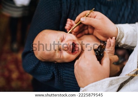 A Christian priest in a church conducts a sacred rite, the ritual of anointing the head of a newborn crying child. Close-up photography, religion.
