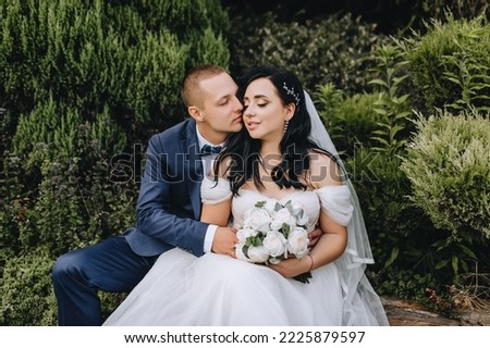 A stylish, young groom in a blue suit and a beautiful, curly brunette bride in a white lace dress gently hug while sitting in a park outdoors. Wedding photography, portrait of newlyweds in love.