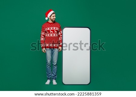 Full body fun merry young man wear red knitted sweater Santa hat posing look at big huge blank screen mobile cell phone with area isolated on plain dark green background New Year 2023 holiday concept