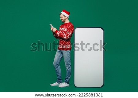 Full body merry young man wear red knitted sweater Santa hat posing huge blank screen mobile cell phone with area use smartphone isolated on plain dark green background New Year 2023 holiday concept