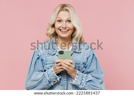 Elderly surprised amazed satisfied happy woman 50s wear denim jacket hold in hand use mobile cell phone isolated on plain pastel light pink background studio portrait. People lifestyle fashion concept