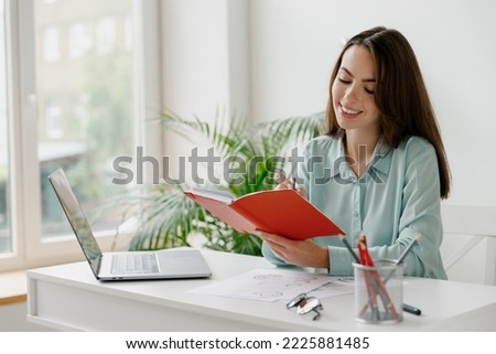 Young smiling happy successful employee business woman 20s in blue shirt writes in notebook sit work at workplace white desk with laptop pc computer at modern office indoors Achievement career concept