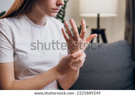 Unhealthy upset young caucasian woman with wrist pain tries to relieve suffering sitting on sofa in living room at home. Close-up of wrist female hand with arthritic joints. Wrist pain concept.