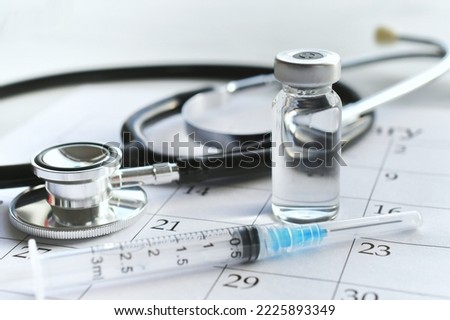 A medical syringe needle with plain unmarked vial of medicine laying on a calendar page with a stethoscope. Concept for scheduling childhood immunizations or covid-19 tetanus or flu shot boosters.  Royalty-Free Stock Photo #2225893349