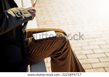 Close-up of a blind man with a walking stick sits on a bench uses a smartphone