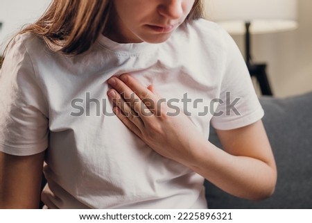 Young unhealthy female sit on couch having difficulty breathing pain of heart, touches his chest with hand. Trouble breathing, chest pain. Heart attack, thoracic osteochondrosis, panic attack concept Royalty-Free Stock Photo #2225896321