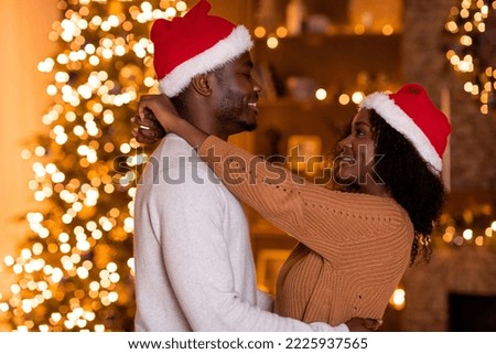 Smiling millennial black guy and female in Santa hats hugging in dark room with Christmas tree with luminous garlands. Celebrate holiday and New Year together at home, love and romance due covid-19