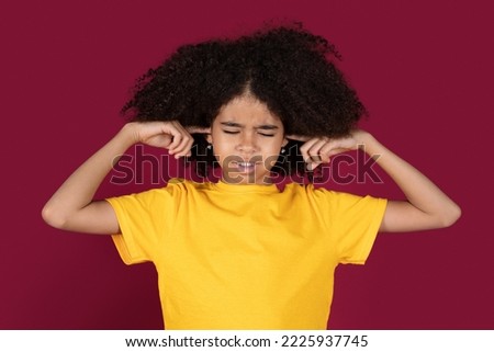 Irritated black girl isolated on burgundy studio background close plug ears with closed eyes, tired by noisy sound, furious angry preteen child avoid ignore unbearable loud noise ask quiet