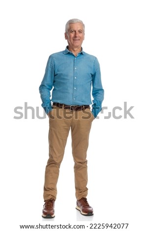 full length picture of happy old man holding hands in pockets and smiling while posing on white background in studio