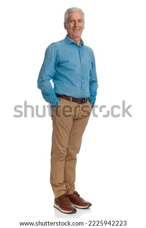 full body picture of happy man in denim shirt holding hands in pockets and smiling while posing on white background in studio