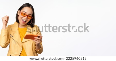 Stylish korean girl in sunglasses, playing mobile video game, laughing and smiling while using smartphone, standing over white background.