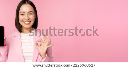 Satisfied smiling asian businesswoman recommending mobile phone app, website company on smartphone, showing screen and okay sign, pink background.