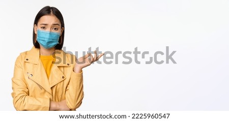 Confused asian woman in medical face mask, looking clueless, puzzled, wearing medical face mask, standing over white studio background.