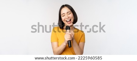 Happy asian girl singing and having fun, holding microphone at karaoke, standing in yellow tshirt against white background. Copy space