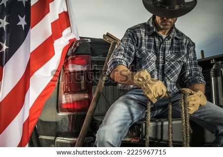 Caucasian Cowboy Rancher in His 40s Seating on His Pickup Truck Cargo Bed with a Rope in His Hands. Preparing Tools For Some Ranch Job. American Flag Next to Him.