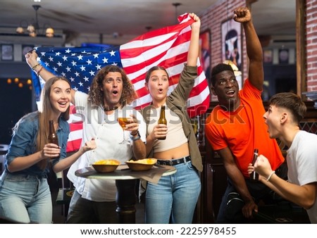 Group of emotional young adults, American football fans cheering for favorite team together in sports bar, waving flag of United States while watching match on TV