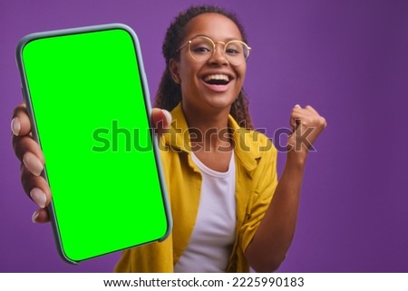 Young overjoyed African American woman holds out phone to camera and makes victorious wave hand wanting to show off achievements in Internet blogging stands in purple studio. Green screen smartphone