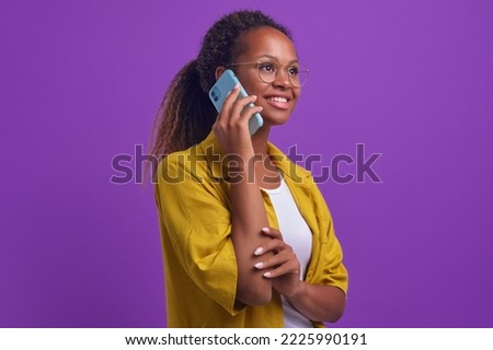 Young charismatic curly African American woman wearing glasses puts mobile phone to ear to make important call and looks into distance smiling broadly stands on lilac background. Copy space Royalty-Free Stock Photo #2225990191