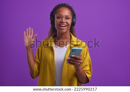Young positive African American woman in wireless headphones uses mobile phone to watch movies and series and waves hand looking at camera stands on isolated lilac background. Musical apps concept
