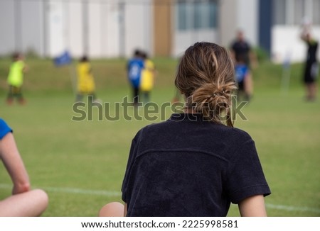 Mom sitting and watching her son playing football in a school football tournament on a sideline with a sunny day. Sport, outdoor active, lifestyle, happy family and soccer mom and soccer dad concept. Royalty-Free Stock Photo #2225998581