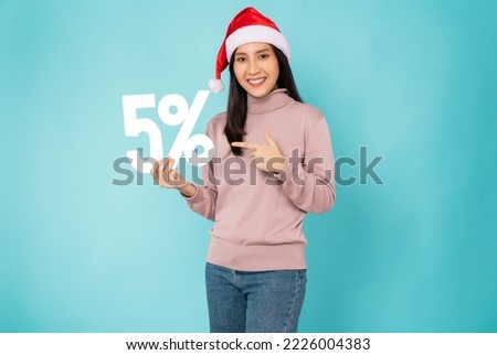 Cheerful beautiful Asian woman wearing christmas hat with with pointing finger on hand holding 5% on light blue background.