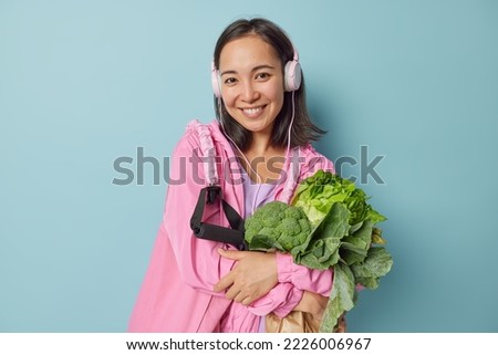 Sport and healthy food concept. Horizontal shot of cheerful Asian woman carries fresh vegetables expander smiles positively returns after training isolated over blue background leads active lifestyle
