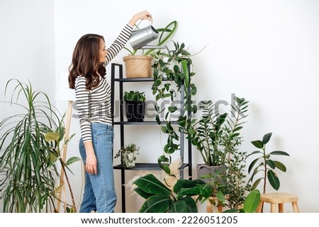 Young woman watering house plants on the rack. At home garden in the corner of a living room with white walls.