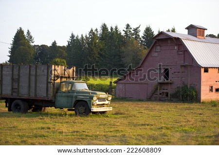 Old rusty truck with a trailer in a field near a large red wooden barn, behind which is growing ever-green trees ryaz. Truck and the house lit up the warm rays of the evening sun. Retro truck.