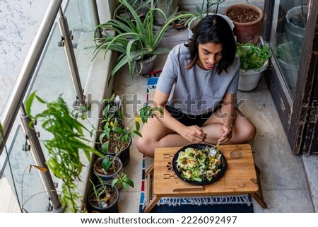 A young Indian woman, dressed in casual clothes, eats a healthy meal while seated in her outdoor balcony surrounded by potted plants and greenery on a summer morning in New Delhi. Royalty-Free Stock Photo #2226092497