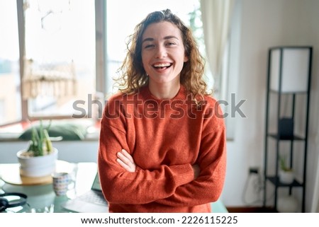 Young smiling pretty lady standing at home looking at camera, happy beautiful positive cheerful professional woman laughing in modern cozy apartment living room or in office, portrait.