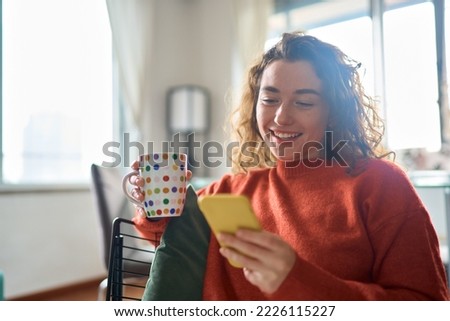 Young happy woman using cell phone drinking coffee relaxing at home. Smiling pretty lady holding smartphone enjoying mobile shopping, checking social media messages, playing game or buying online. Royalty-Free Stock Photo #2226115227