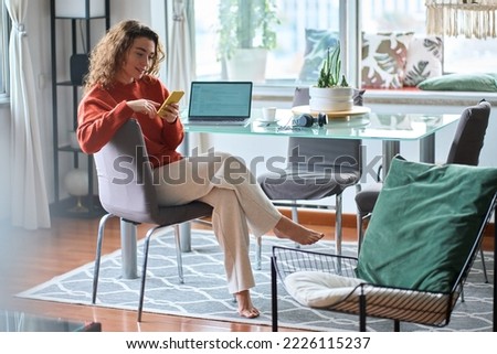 Young smiling pretty relaxed woman sitting on chair holding smartphone using cellphone modern technology, looking at mobile phone while remote working at table or e learning at cozy home interior.