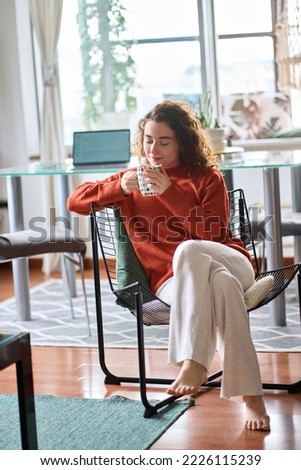 Young adult smiling pretty woman sitting on chair holding cup drinking tea or coffee relaxing at home. Happy calm lady enjoying warm hot drink with mug in hands daydreaming in cozy morning. Vertical