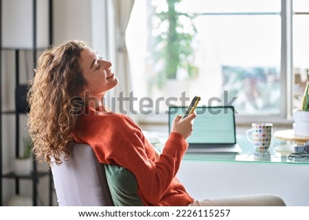 Young smiling pretty woman holding smartphone using cell mobile phone taking break relaxing while remote working or learning from home sitting on chair at table with laptop. Royalty-Free Stock Photo #2226115257