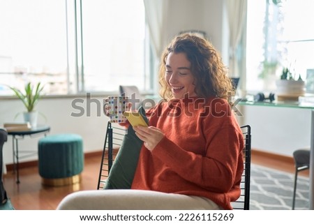 Young smiling pretty woman holding smartphone technology drinking tea or coffee relaxing at home. Happy positive lady enjoying using mobile cell phone having fun while buying in ecommerce shop.