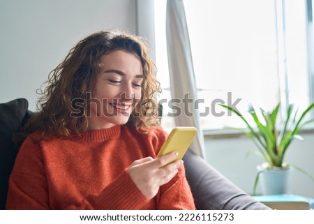Smiling young woman sitting on couch using cell phone, happy lady holding smartphone, looking at cellphone doing online shopping in mobile apps ordering ecommerce products or checking social media.