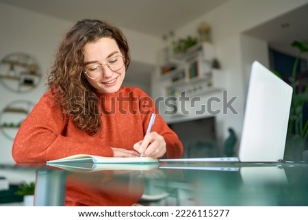 Young smiling pretty woman student using laptop elearning or remote working at home office using laptop computer watching webinar, learning web course, studying online sitting at table, writing notes. Royalty-Free Stock Photo #2226115277