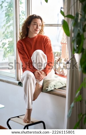 Happy serene young woman sitting on windowsill relaxing at home looking through window. Smiling calm lady chilling in apartment, dreaming, thinking of peaceful time enjoying peace of mind. Vertical