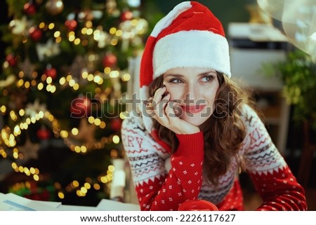 Christmas time. pensive modern middle aged small business owner woman in santa hat in modern green office with Christmas tree in red Christmas sweater.
