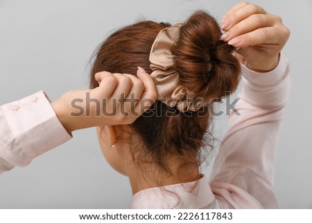 Woman with bun hairstyle adjusting silk scrunchy on grey background