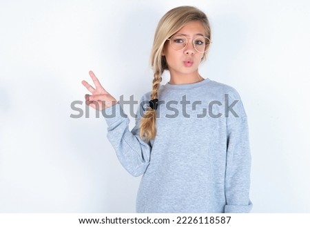 caucasian teen girl wearing gray sweater over white background makes peace gesture keeps lips folded shows v sign. Body language concept