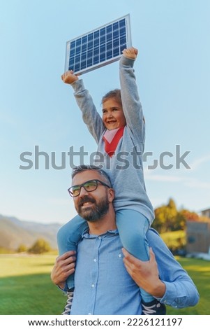 Father with his little daughter on piggyback, catching sun at solar panel,charging it at their backyard. Alternative energy, saving resources and sustainable lifestyle concept. Royalty-Free Stock Photo #2226121197